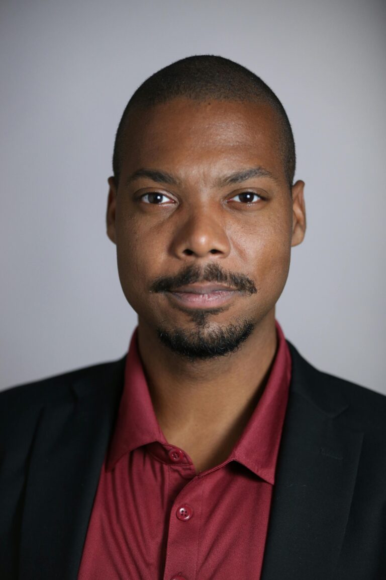 Portrait of a confident black man in formal attire against a gray background.