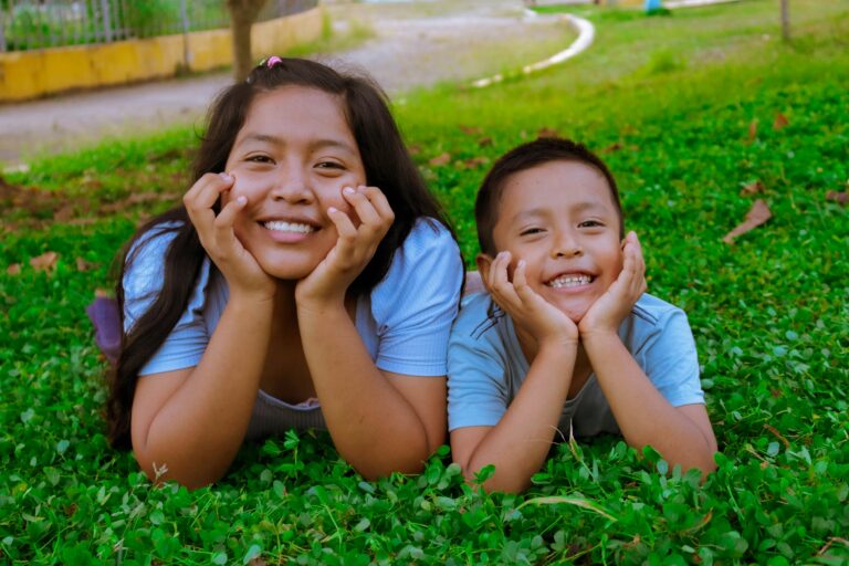 A cheerful duo of a teenager and a child laying on grass, sharing joyful smiles outdoors.